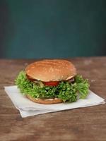 Big Tasty beef patty Burger rests on craft white paper against wooden table. A Juicy green Salad leaf and a red Tomato lie near the Burger. Vertical shot photo