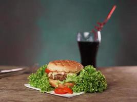 un sabroso hamburguesa mentiras en un de madera mesa en contra un verde antecedentes. allí es un vaso de soda, un cuchillo y un tenedor en el mesa foto