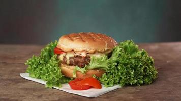 Big Tasty beef patty Burger rests on craft white paper against wooden table. A Juicy green Salad leaf and a red Tomato lie near the Burger photo