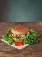 Big Burger lies on craft white paper against wooden table. A Juicy green Salad leaf and a red Tomato lie near the Burger. Vertical shot photo