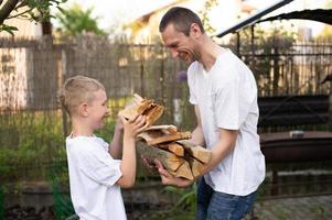 A cute boy in a white T-shirt holds firewood and gives it to his dad. photo