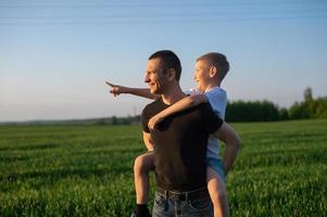The boy is sitting on his dad's back pointing his finger to the side and they laugh photo