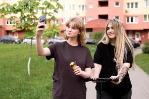 Two funny girls are sitting on a swing in the city and taking selfies on their phone. Teenagers smile photo