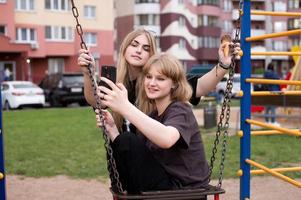 Two funny girls are sitting on a swing in the city and taking selfies on their phone. Teenagers smile photo