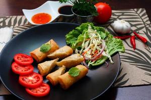 Fried spring rolls with vegetables and tomatoes placed in a black plate on a black wooden table and dipping sauce. photo