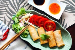 Fried spring rolls, vegetables and tomatoes placed in a green leaf shape plate on a black wooden table and dipping sauce. photo