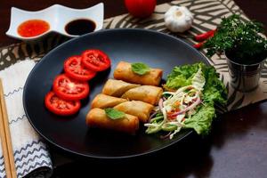 Fried spring rolls with vegetables and tomatoes placed in a black plate on a black wooden table and dipping sauce. photo