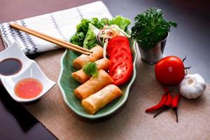Fried spring rolls, vegetables and tomatoes placed in a green leaf shape plate on a black wooden table and dipping sauce. photo