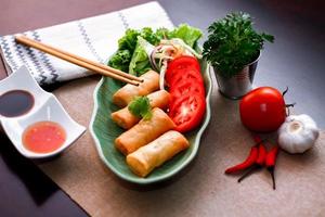 Fried spring rolls, vegetables and tomatoes placed in a green leaf shape plate on a black wooden table and dipping sauce. photo