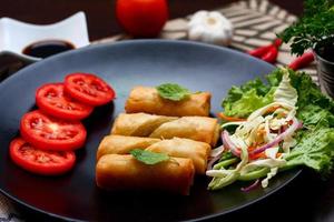 frito primavera rollos con vegetales y Tomates metido en un negro plato en un negro de madera mesa y inmersión salsa. foto