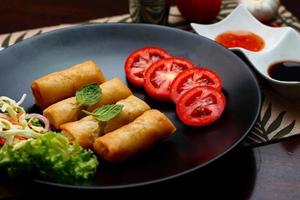 Fried spring rolls with vegetables and tomatoes placed in a black plate on a black wooden table and dipping sauce. photo