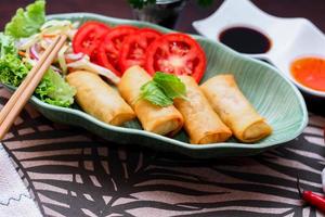 Fried spring rolls, vegetables and tomatoes placed in a green leaf shape plate on a black wooden table and dipping sauce. photo