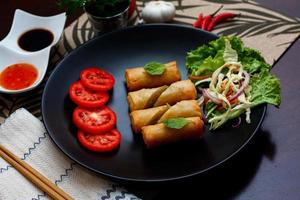 Fried spring rolls with vegetables and tomatoes placed in a black plate on a black wooden table and dipping sauce. photo