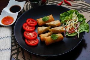 Fried spring rolls with vegetables and tomatoes placed in a black plate on a black wooden table and dipping sauce. photo