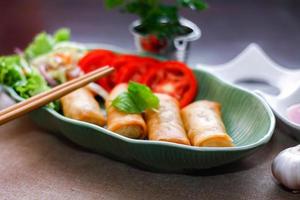 Fried spring rolls, vegetables and tomatoes placed in a green leaf shape plate on a black wooden table and dipping sauce. photo