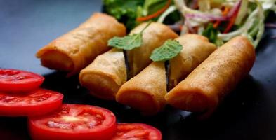 frito primavera rollos con vegetales y Tomates metido en un negro plato en un negro de madera mesa y inmersión salsa. foto