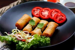 Fried spring rolls with vegetables and tomatoes placed in a black plate on a black wooden table and dipping sauce. photo