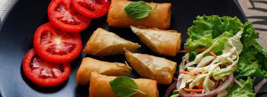 Fried spring rolls with vegetables and tomatoes placed in a black plate on a black wooden table and dipping sauce. photo