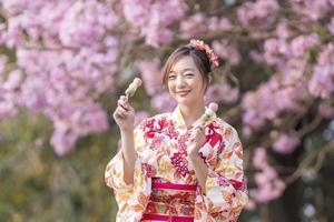 japonés mujer en tradicional kimono vestir participación dulce hanami dango postre mientras caminando en el parque a Cereza florecer árbol durante primavera sakura festival con Copiar espacio foto