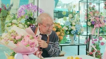 comércio eletrônico negócios, 1 Senior masculino florista trabalhadores demonstrar e mostrar floral arranjos através da conectados viver transmissão com Smartphone inscrição dentro uma brilhante flor comprar, uma lindo flores loja. video