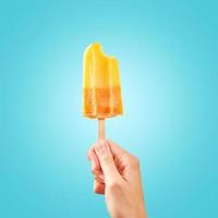 Bitten red frozen fruit ice cream popsicle in woman hand on blue background photo