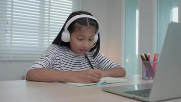 online tutor online course for Asian children. video conference on laptop at home. play back online course, girl student wear earphones to study, notes homework, video classes, new education.