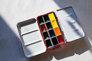 Photo of watercolor paints in cuvettes, a metal case for watercolor painting on watercolor white paper.