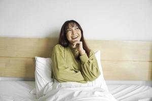 A thoughtful young Asian woman is looking aside and holding her chin while sitting on the bed photo