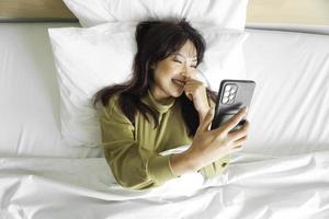 un sonriente joven asiático mujer es riendo mientras participación su teléfono y acostado en el cama foto