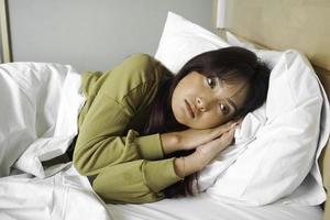 Thoughtful Asian woman looks pensively above dressed in sweatshirt poses thinks about future while lying on the bed photo