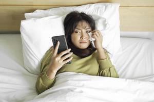 A sad young Asian woman is crying over her phone while lying on the bed photo