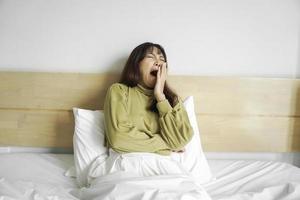 Cute Asian woman stretching out and yawning while sitting on her bed in bright bedroom photo