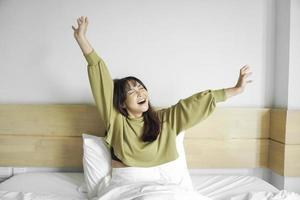 retrato de belleza contento asiático mujer despertando, despertar arriba en el cama. joven hermosa asiático niña extensión su brazos en Fresco temprano mañana, relajarse pacífico Mañana rutina bostezo concepto bandera foto