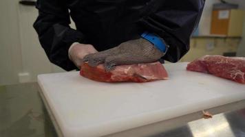 Butcher in metal protective gloves Cuts a freshness raw piece of Beef Meat on a cutting white board with a Knife. Close up video