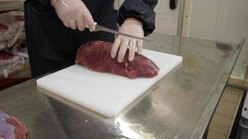 een slager vervelend wit beschermend handschoenen zet een groot stuk van vers rauw rundvlees vlees Aan een snijdend bord. arbeider bezuinigingen rauw vlees met scherp mes in steaks video