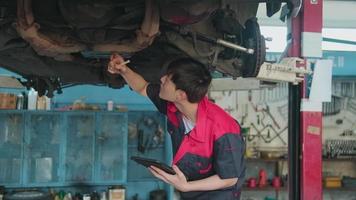 Professional young Asian male motor mechanic inspects undercarriage of electric car lifted by forklift jack for repair at garage, automotive maintenance service works industry occupation business. video