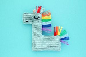 Crochet amigurumi handmade stuffed soft blue unicorn toy with rainbow mane on blue background. Handwork, hobby. Craft diy newborn pregnancy concept. Knitted doll for little baby. Close up flat lay photo