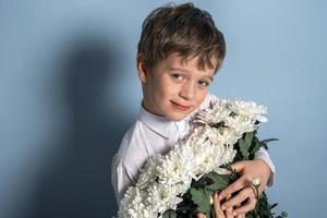 A cute Caucasian boy in a white shirt holds a bouquet of white flowers in his hands and smiles. photo