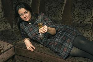 A young woman in a plaid dress drinks wine from a glass lying on a home sofa photo