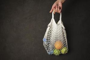 A woman's hand holds a string bag with a bottle of water, an orange and green apples photo