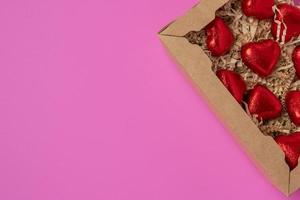 A group of heart-shaped chocolates wrapped in red foil, lying in a gift box on a pink background photo