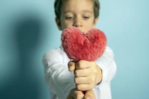 un caucásico chico en un blanco camisa sostiene un mullido rojo corazón en su manos. foto