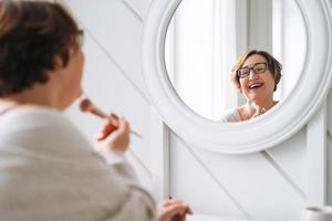 sonriente medio Envejecido más Talla mujer haciendo maquillaje cerca espejo a hogar foto