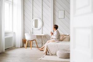 Middle aged plus size woman with brunette curly hair with cup of tea in hands sitting on bed at home photo