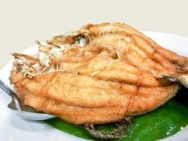 frito pargo pescado coronado con pescado salsa en plátano hoja y blanco cerámico plato con inoxidable acero cuchara., esta comida es más popular tailandés comida en el restaurante. foto