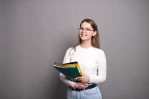 linda estudiante con lentes sostiene vistoso carpetas en un gris antecedentes foto