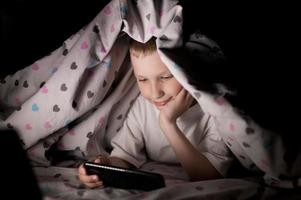 A little boy lies under a blanket at night playing on the phone photo