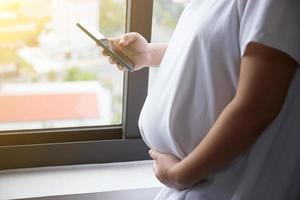 joven asiático embarazada mujer utilizando inteligente teléfono para comprobación Noticias mientras en pie cerca el ventana foto