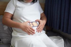 Image of Asian pregnant smiling woman sitting and posing while showing ultrasound scans, look into the camera photo