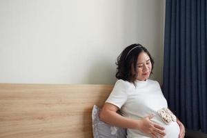 foto de contento asiático embarazada mujer sentado en cama y conmovedor su barriga a hogar . con pequeño Zapatos para el no nacido bebé en el barriga